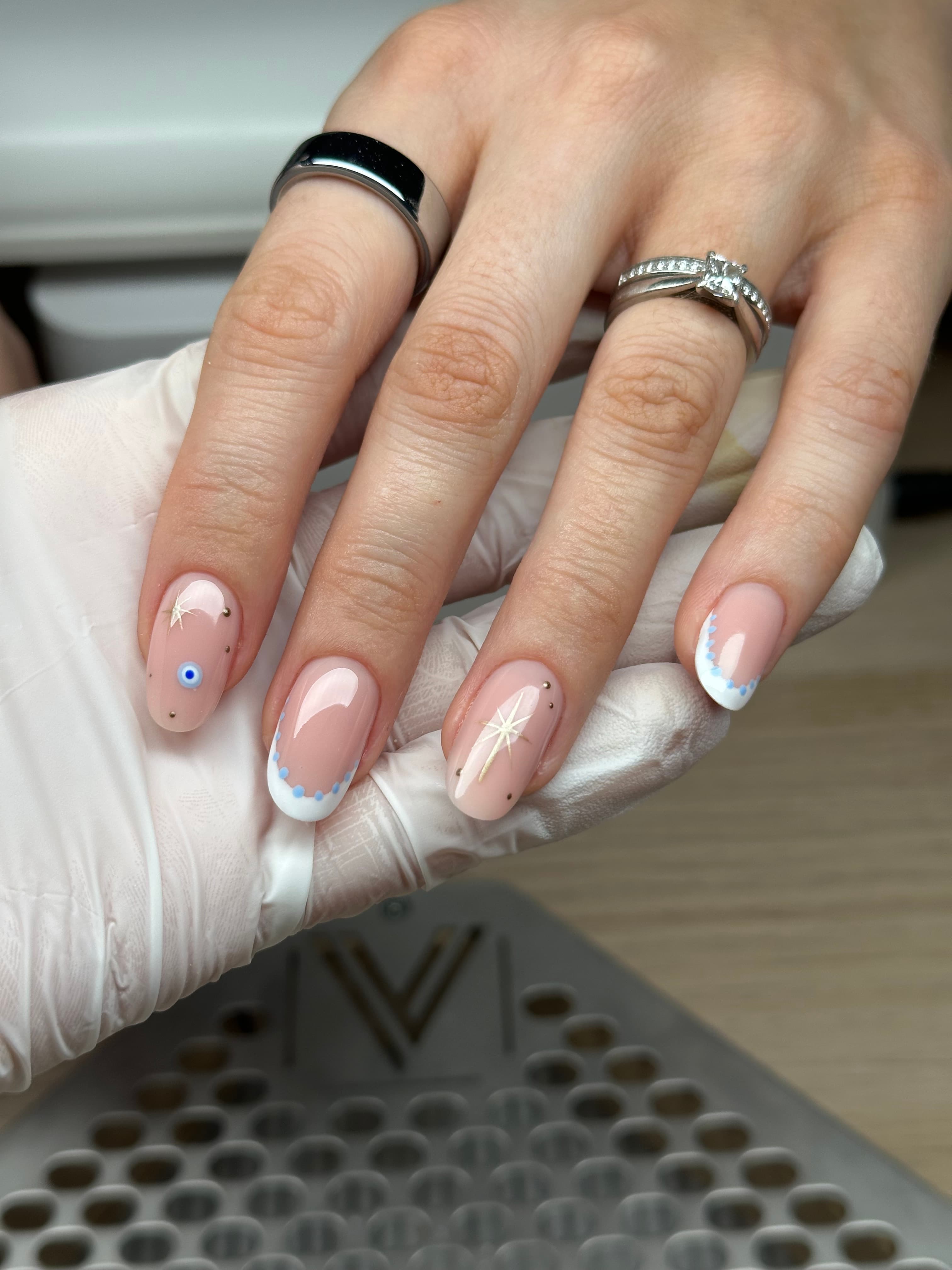 Oval nude nails with white French tips lined in blue dots, gold star accents, and an evil eye charm nail